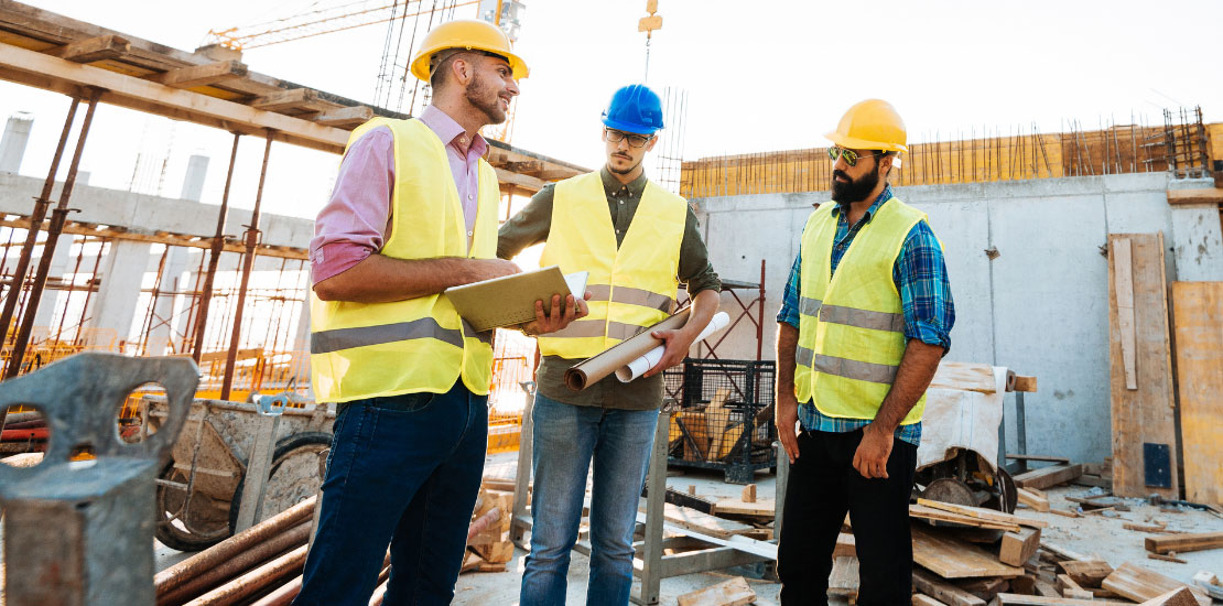 Three construction workers in high visibility vests and safety helmets standing at a construction site with building materials and scaffolding in the background.