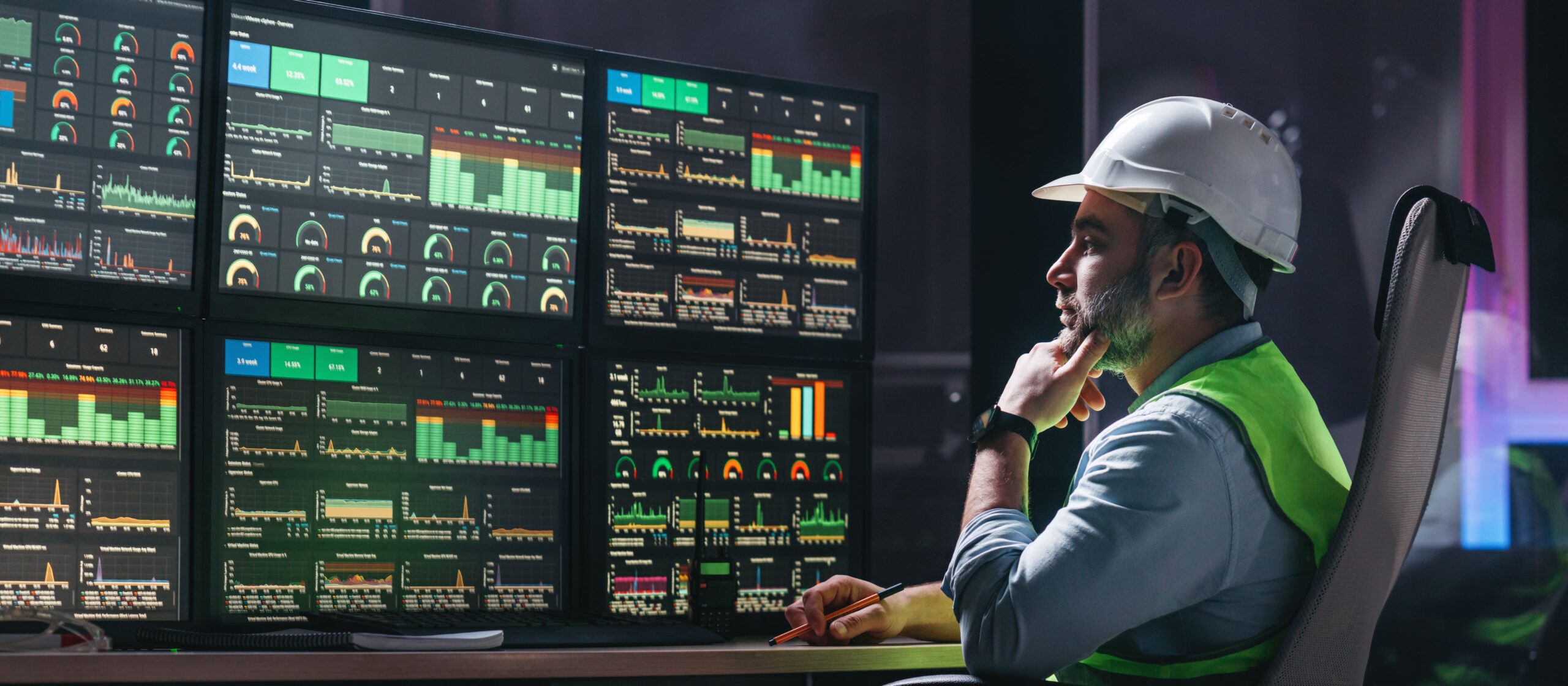 A man in a hard hat and reflective vest analyzing data on multiple computer screens in a control room