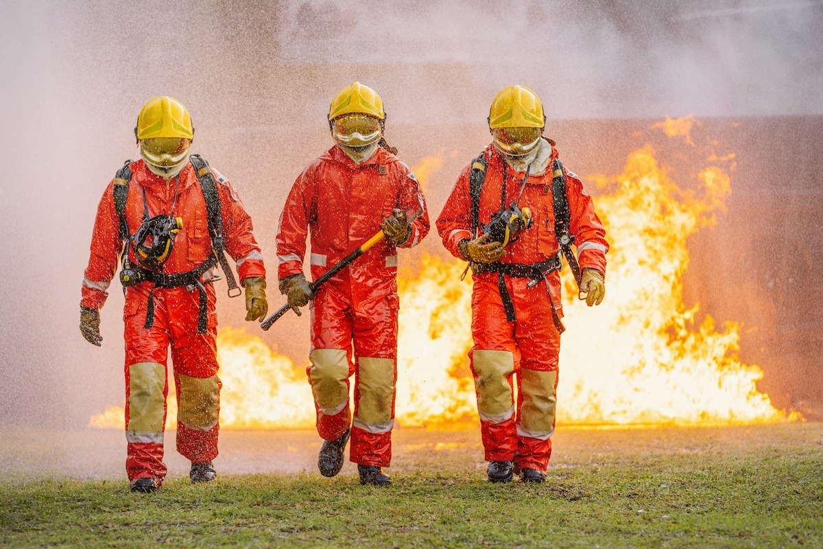 Three firefighters are walking through fire, one of them holding hose